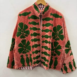 May include: Coral-colored jacket with green embroidered floral and abstract designs. The jacket has a mandarin collar and long sleeves. The interior lining features a red and white striped pattern. The jacket is hanging on a wooden hanger.