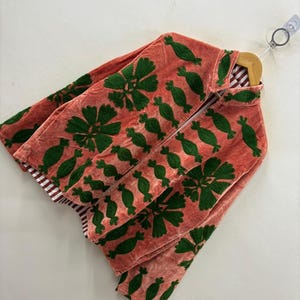 May include: A coral-colored jacket with green embroidered floral and pod-like designs. The jacket is made of a soft, textured fabric and features a striped lining. It is hanging on a wooden hanger against a white background.