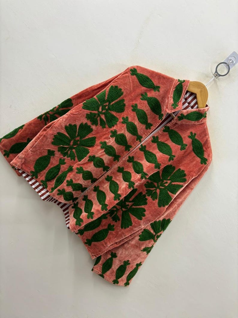 May include: A coral-colored jacket with green embroidered floral and pod-like designs. The jacket is made of a soft, textured fabric and features a striped lining. It is hanging on a wooden hanger against a white background.