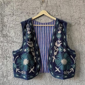 May include: A navy blue embroidered vest with teal and white floral designs. The vest has a blue and white striped lining and is suspended on a wooden hanger. The embroidery includes large flowers and scrolling vines.