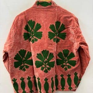 May include: A coral-colored jacket with green floral embroidery. The jacket features large, stylized flower designs on the back and smaller, decorative elements along the bottom hem and collar. The fabric has a soft, textured appearance.