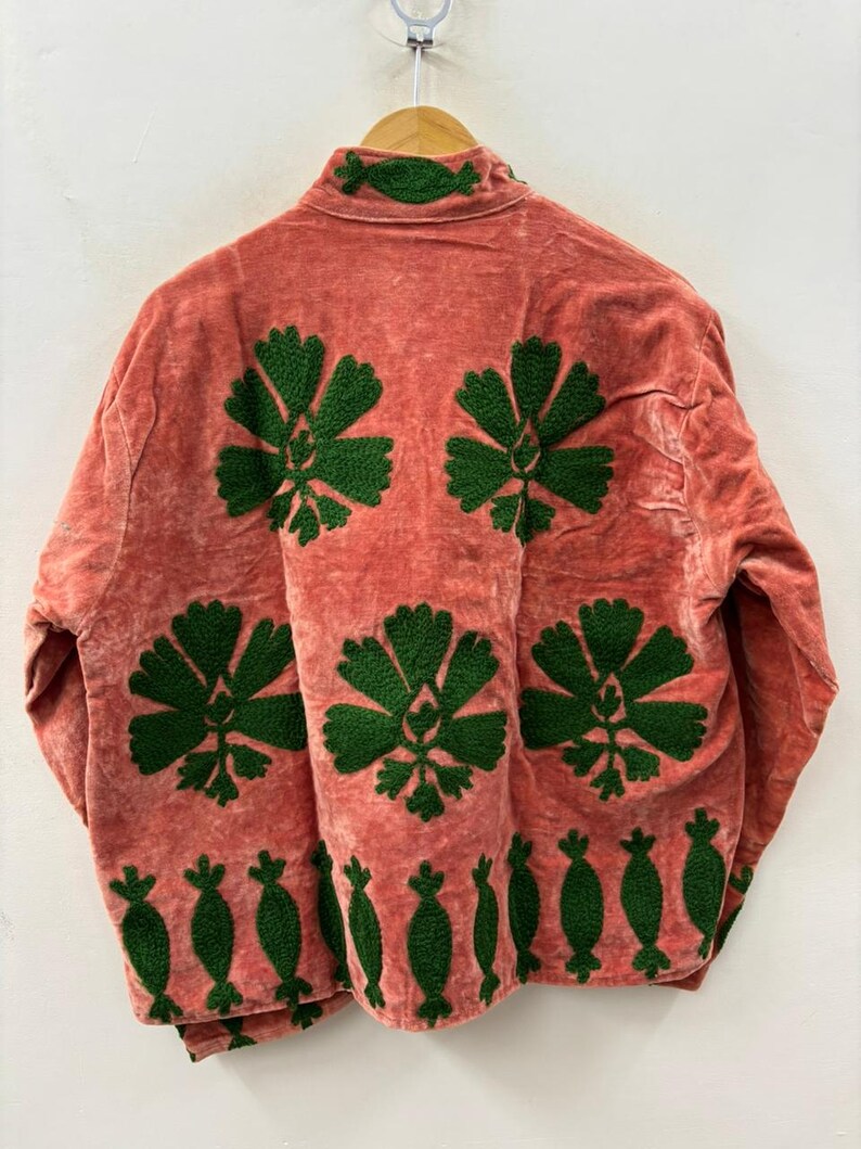 May include: A coral-colored jacket with green floral embroidery. The jacket features large, stylized flower designs on the back and smaller, decorative elements along the bottom hem and collar. The fabric has a soft, textured appearance.