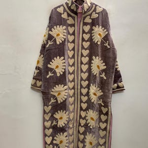May include: A long, mauve-colored velvet coat featuring cream-colored floral embroidery and a vertical pattern of hearts along the front opening and collar. The garment has long sleeves, a high neckline, and side slits, with a light pink striped lining.