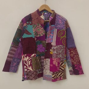 May include: A patchwork jacket in shades of purple, burgundy, and teal. The jacket features a mandarin collar and is composed of various fabric squares with floral and geometric patterns. It is hanging on a wooden hanger.