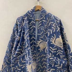 Könnte beinhalten: Eine blaue Samtjacke mit einem aufwendigen, cremefarbenen Blumen- und Rankenstickmuster. Das Kleidungsstück hat einen breiten Schalkragen, lange Ärmel und zwei Vordertaschen. Das Innenfutter ist sichtbar und zeigt einen blau-weiß gestreiften Stoff.