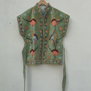 May include: An olive green embroidered jacket with a mandarin collar and no sleeves. The jacket features a floral design in peach, blue, and green. It has a tie closure and is hanging on a wooden hanger.
