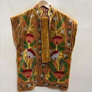 May include: A mustard yellow sleeveless waistcoat featuring intricate floral embroidery in shades of red, white, and green. The garment has a high collar, a front tie closure, and a vertical striped lining visible at the neck and arm openings.