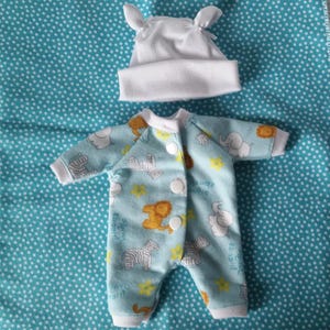 May include: A light blue long-sleeved onesie featuring a repeating pattern of white elephants, zebras, and orange lions with yellow stars, secured with three white snaps, paired with a solid white beanie hat with two small rounded ears on top.