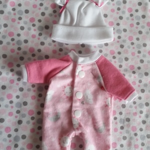 May include: A white beanie with small pink bows and a pink and white long-sleeved romper featuring a pattern of suns and clouds. The romper has a white collar, white sleeve cuffs, and three white snap buttons down the front center for closure.