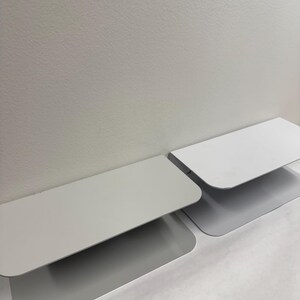 May include: Two modern, minimalist, gray metal shelves with rounded corners are displayed against a white wall. The shelves are rectangular with a slightly curved design, creating a clean, contemporary aesthetic.