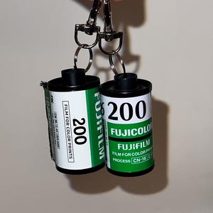Puede incluir: Llavero con dos cartuchos de película Fujifilm 200. Los cartuchos son negros y blancos con etiquetas verdes y blancas. El texto "200" es visible. El llavero tiene cierres y anillos plateados.