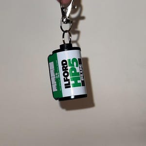 May include: A keychain featuring a miniature film roll. The film roll is white and green with the text "ILFORD HP5 PLUS" printed on it. The keychain has a silver clasp and a small ring to attach the film roll.