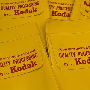 Può includere: Primo piano di diverse buste gialle per l'elaborazione di pellicole Kodak. Le buste hanno una parte superiore curva e presentano il testo "YOUR PICTURES DESERVE QUALITY PROCESSING by... Kodak" in rosso.