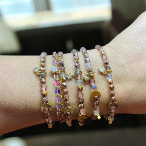 May include: A collection of beaded bracelets in shades of pink, gold, and iridescent colors. Each bracelet features small, decorative charms, including hearts, stars, and half-moon shapes. The bracelets are strung on elastic and designed to be worn stacked.