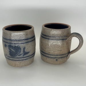 May include: Two barrel-shaped stoneware mugs with a textured beige finish and dark blue horizontal bands. One mug features a dark blue floral design, while the other has a handle and a faint embossed logo on the front. Both have dark interiors.