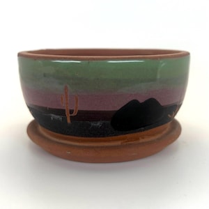 May include: A round terracotta planter with a matching saucer, featuring a horizontal band design in shades of green, mauve, and black. The exterior surface displays a painted desert landscape motif with a small orange cactus and a black mountain.