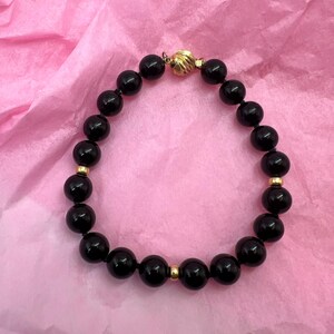 May include: A black beaded bracelet with gold-colored accents. The bracelet features large, round, glossy black beads strung together with small gold-colored beads interspersed throughout. The clasp is a textured gold-colored sphere.