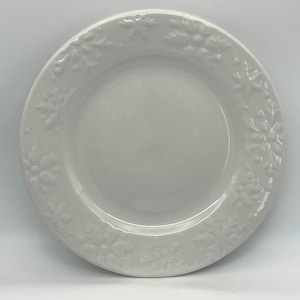 Puede incluir: Un plato de cerámica blanco con un diseño floral en relieve alrededor del borde. El plato tiene un acabado liso y brillante y un color blanco ligeramente apagado. El patrón floral es un diseño repetitivo de flores estilizadas.