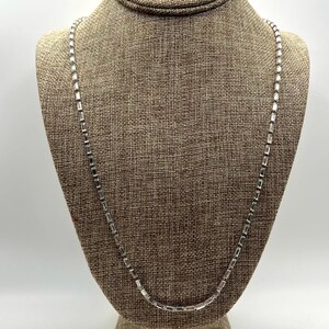 May include: A silver-toned necklace with rectangular links is displayed on a mannequin. The necklace is draped in a gentle curve, showcasing its delicate design. The background is a neutral tan color.