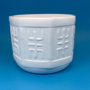 May include: A white ceramic planter with a cylindrical shape and a smooth rim. The exterior features a repeating embossed geometric pattern set against a textured background, framed by vertical and horizontal smooth bands around the body.