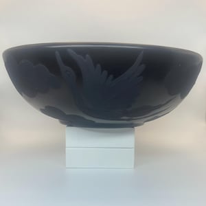 Puede incluir: Un cuenco decorativo azul oscuro con un diseño de pájaro y nubes. El cuenco, de forma ovalada, se apoya sobre una base rectangular blanca. El pájaro está representado en vuelo con las alas extendidas.