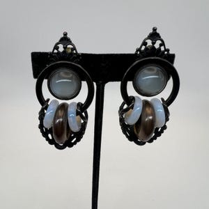 Pode incluir: Um par de brincos de metal preto com um design circular. Cada brinco apresenta uma pedra grande azul clara no centro, rodeada por um anel preto. Abaixo do anel, existem três anéis menores, dois azuis claros e um castanho.
