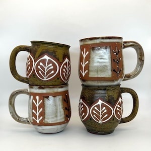 May include: Four ceramic mugs stacked. The mugs have a brown, green, and white leaf design. Each mug has a large handle and a unique leaf pattern. The mugs are on a white background.