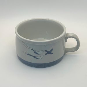 May include: A ceramic soup mug with a light gray body and a blue base. The mug features a curved handle and a design of two stylized blue birds in flight. The mug is suitable for serving soup, coffee, or tea.