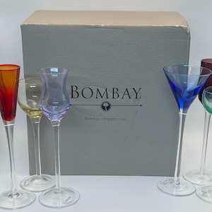 May include: A set of colorful stemmed glasses in various shades, including red, yellow, purple, and blue. The glasses are displayed in front of a gray box with the "BOMBAY" logo. The glasses have clear stems and bases.