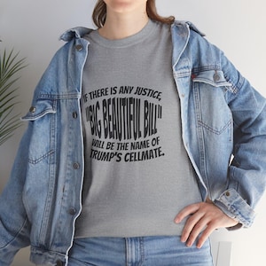May include: A heather gray t-shirt featuring black text that reads: IF THERE IS ANY JUSTICE, "BIG BEAUTIFUL BILL" WILL BE THE NAME OF TRUMP'S CELLMATE. The t-shirt is worn under a light blue denim jacket with metal buttons and blue denim jeans.
