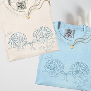 May include: A cream t-shirt and a light blue t-shirt, each featuring a dark blue line art graphic of two seashells connected by a bow. The cream shirt has the text The Bride, and the blue shirt has the text The Bach. Both include Amelia's Bachelorette Miami 2024.