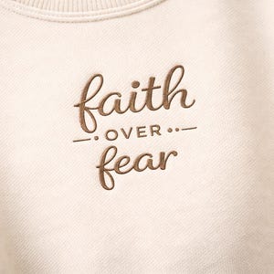 Puede incluir: Sudadera de color crema con la frase bordada "faith over fear" en fuente cursiva marr&oacute;n. El texto est&aacute; centrado e incluye una l&iacute;nea horizontal y puntos entre las palabras. La sudadera tiene un cuello acanalado.