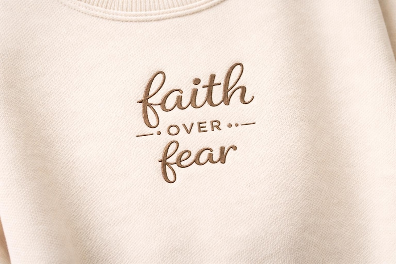 Puede incluir: Sudadera de color crema con la frase bordada "faith over fear" en fuente cursiva marr&oacute;n. El texto est&aacute; centrado e incluye una l&iacute;nea horizontal y puntos entre las palabras. La sudadera tiene un cuello acanalado.