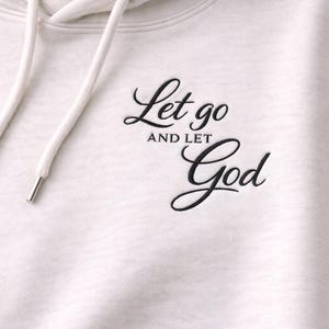 May include: Cream-colored hoodie with a drawstring and metal eyelets. The phrase "Let go and let God" is embroidered in black script on the front. The hoodie has a hood and a soft, textured appearance.