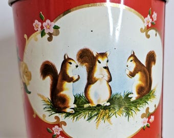 Può includere: Un contenitore in metallo rosso vintage con un design decorativo. Il contenitore presenta un pannello ovale centrale con tre scoiattoli dei cartoni animati. I bordi superiore e inferiore hanno volute dorate. Sono presenti anche fiori rosa e fogliame verde.