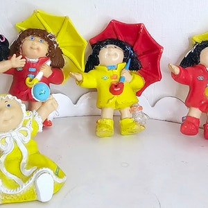 Puede incluir: Una colección de ocho figuras de Cabbage Patch Kids. Las figuras están vestidas con diferentes atuendos, incluyendo un uniforme de fútbol, un vestido amarillo, un impermeable rojo y un mono azul. Algunas de las figuras sostienen accesorios, como un balón de fútbol, un biberón y un paraguas.