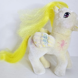 Könnte beinhalten: Ein gelbes und weißes Plüschpony mit gelber Mähne und Schweif. Das Pony hat ein blaues und grünes Blumenmuster auf dem Rücken und ein pinkfarbenes Blumenmuster an der Seite.
