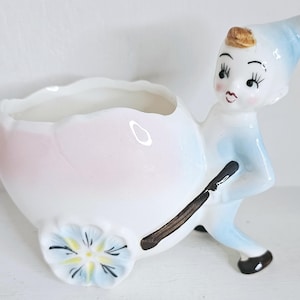 May include: A vintage ceramic planter featuring an elf pushing a wheelbarrow. The planter is white with pink and blue details, and a blue and yellow flower on the wheel. The elf wears a blue hat and holds a brown handle.