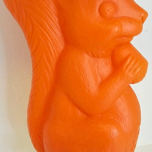 May include: Orange plastic squirrel figurine holding a nut in its paws. The squirrel is sitting on a white base.