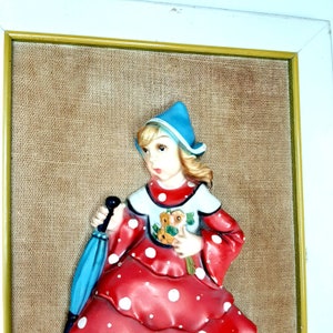May include: A framed 3D wall art piece featuring a girl in a red and white polka dot dress, blue hat, and holding a blue umbrella. The background is a brown burlap fabric.