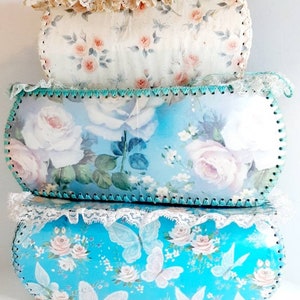 May include: Three vintage storage boxes with floral patterns. The top box is white with pink roses, the middle box is blue with white roses, and the bottom box is blue with white butterflies and pink roses.