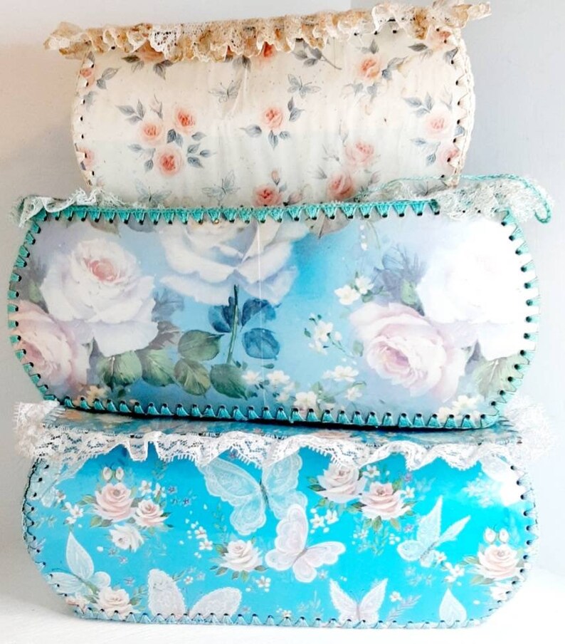 May include: Three vintage storage boxes with floral patterns. The top box is white with pink roses, the middle box is blue with white roses, and the bottom box is blue with white butterflies and pink roses.