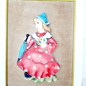 May include: A framed wall decor featuring a 3D ceramic figurine of a girl wearing a red dress with white polka dots, a blue hat, and holding a bouquet of flowers. The background is a beige fabric.