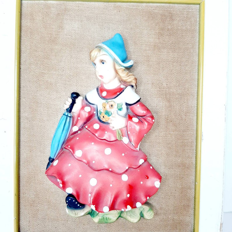 May include: A framed wall decor featuring a 3D ceramic figurine of a girl wearing a red dress with white polka dots, a blue hat, and holding a bouquet of flowers. The background is a beige fabric.