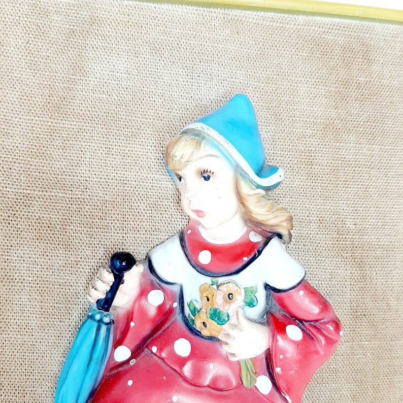 May include: A vintage figurine of a girl wearing a red and white polka dot dress, a blue hat, and holding a bouquet of yellow flowers. She is holding a blue umbrella in her left hand.