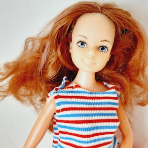 May include: A red and blue striped shirt with a white background. The shirt is worn by a doll with long, curly red hair and blue eyes.