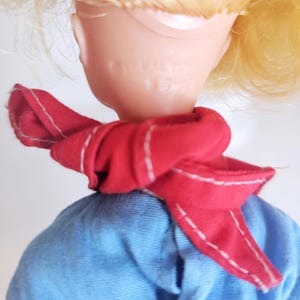 K&ouml;nnte beinhalten: Ein Vintage-Puppenkopf mit blonden Haaren und einem roten Schal mit wei&szlig;en N&auml;hten. Die Puppe tr&auml;gt ein blaues Hemd. Auf der R&uuml;ckseite des Puppenkopfes steht der Text "Ideal Toy" und "9-2".