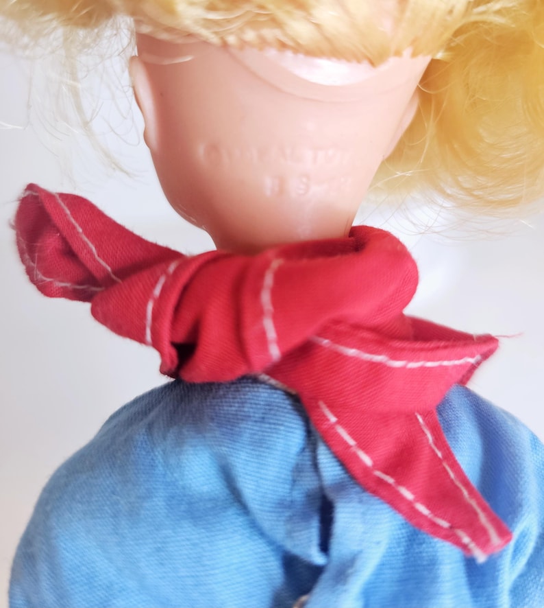 K&ouml;nnte beinhalten: Ein Vintage-Puppenkopf mit blonden Haaren und einem roten Schal mit wei&szlig;en N&auml;hten. Die Puppe tr&auml;gt ein blaues Hemd. Auf der R&uuml;ckseite des Puppenkopfes steht der Text "Ideal Toy" und "9-2".