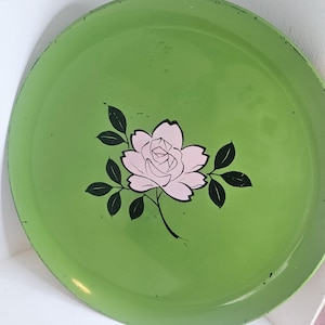 May include: Circular, vintage green metal tray featuring a pink rose and black leaf design. The tray has a slightly worn look, enhancing its retro appeal. A decorative item suitable for serving or display.