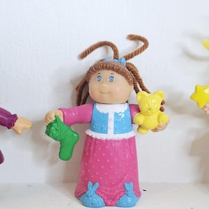 May include: Three Cabbage Patch Kids dolls. The dolls have yarn hair and are wearing dresses and holding accessories. One doll wears a purple dress and holds a green glove. Another doll wears a pink dress and holds a green sock and a yellow teddy bear. The third doll wears a purple leotard and tutu and holds a yellow star wand.